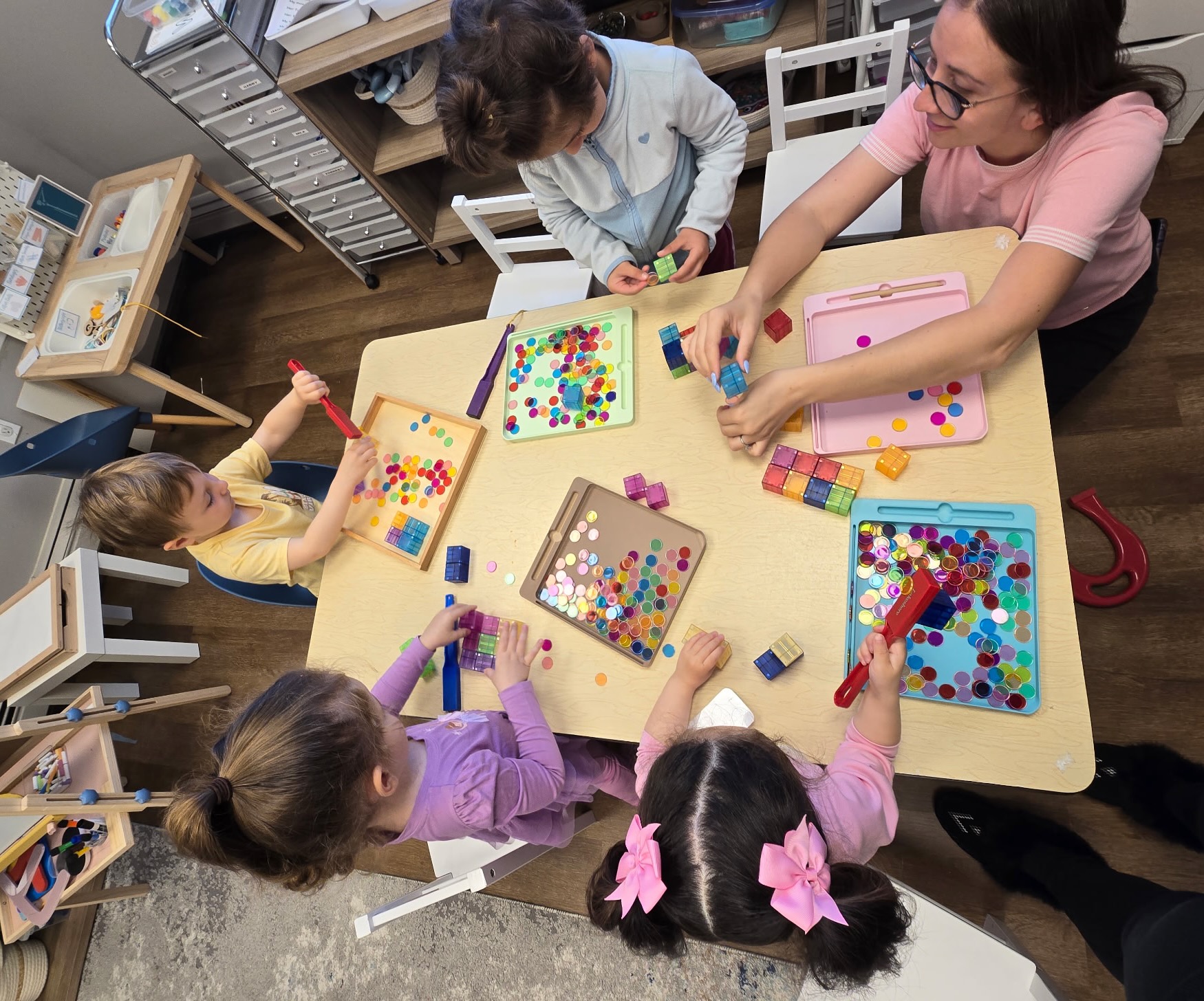 Children and a guide exploring colorful translucent counters and magnetic tiles at a Montessori-inspired work table